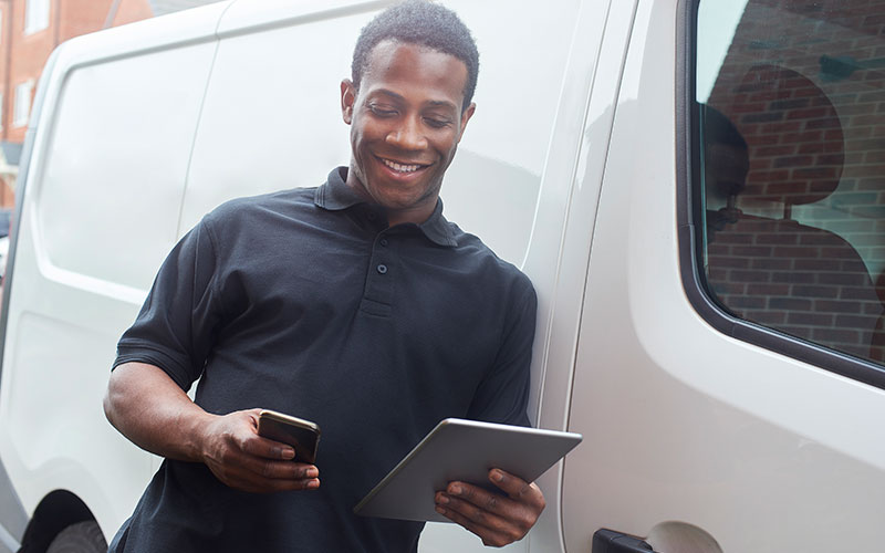 A tradesman stood at the side of his van using a digital tablet and smart phone