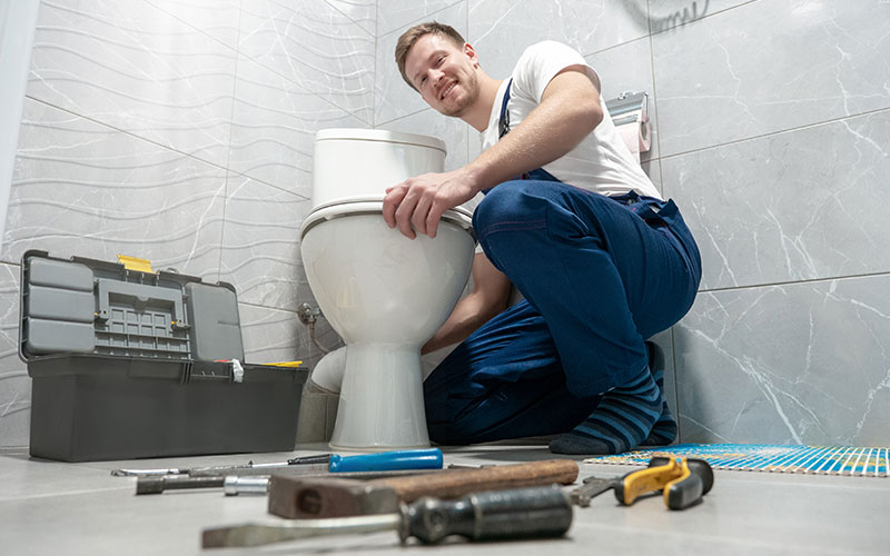 smiling man plumber in uniform repairing toilet bowl using instrument kit looks happy professional repair service.