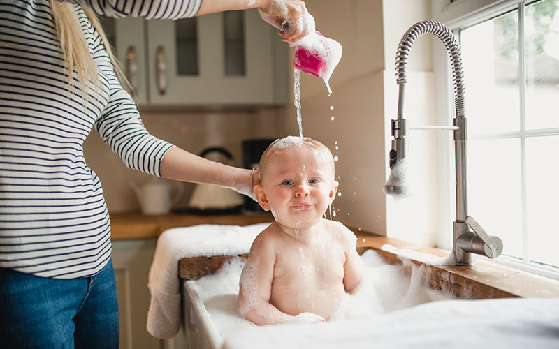 A small baby pulls a silly face in the sink as his mother pours warm water over his head to wash his hair.