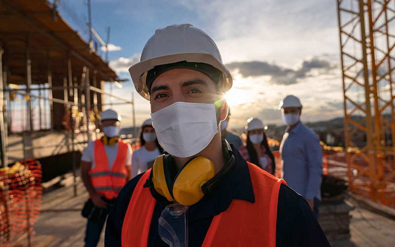 Portrait of a happy construction worker at a building site wearing a facemask with a group of workers at the background